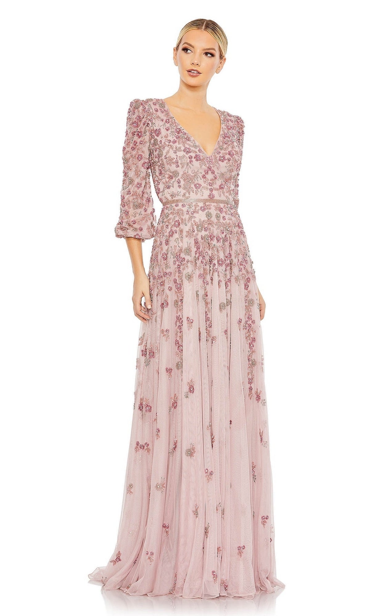 A woman wears the Mac Duggal 5497 Long Sleeve Beaded Formal Dress, a floor-length light pink gown with sheer sleeves and floral embroidery, standing against a plain background.